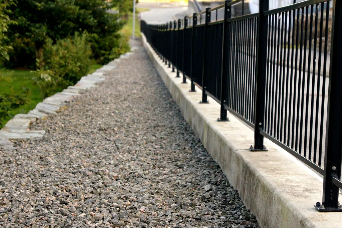 A narrow crushed-stone path held by a stone edge on one side and a curb with a metal railing on the other