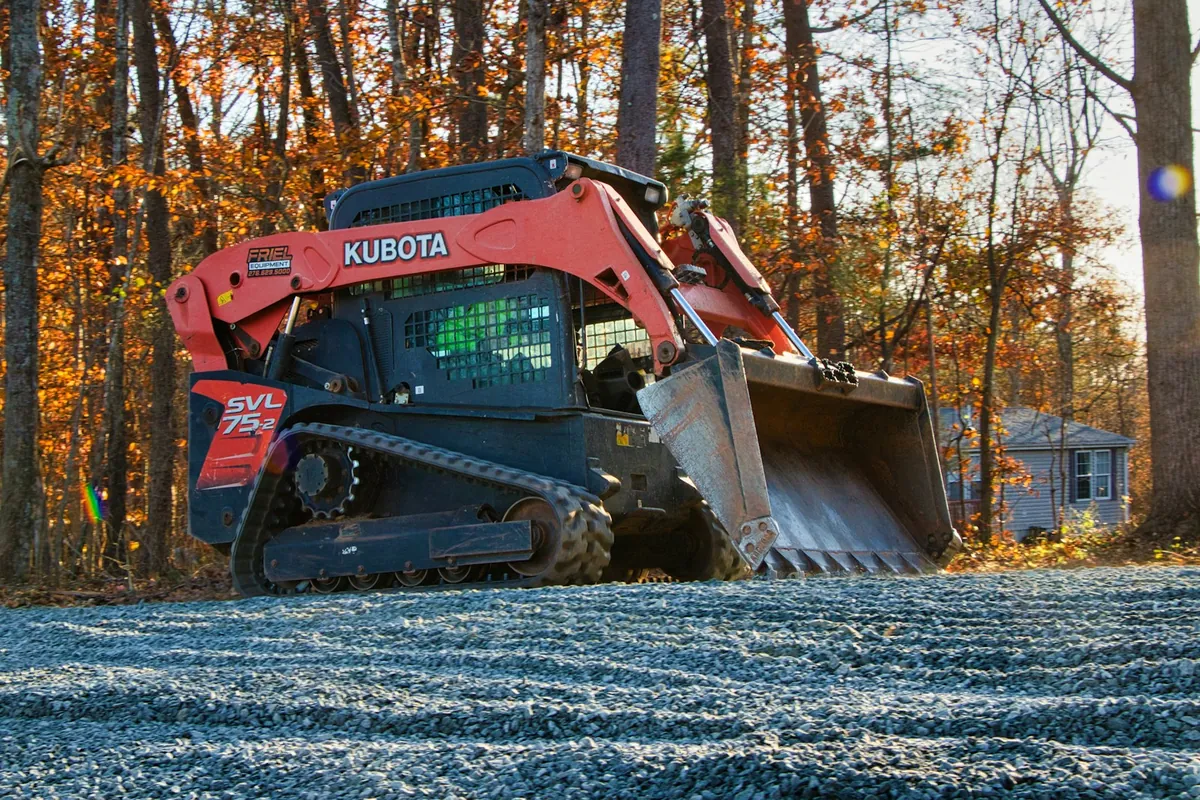 A compact track loader on a fresh spread of crushed gray gravel by a house in the woods in fall