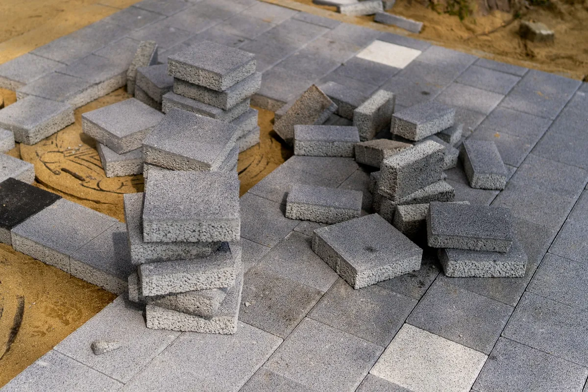 Gray concrete pavers stacked on a sand bed beside a section of patio already laid