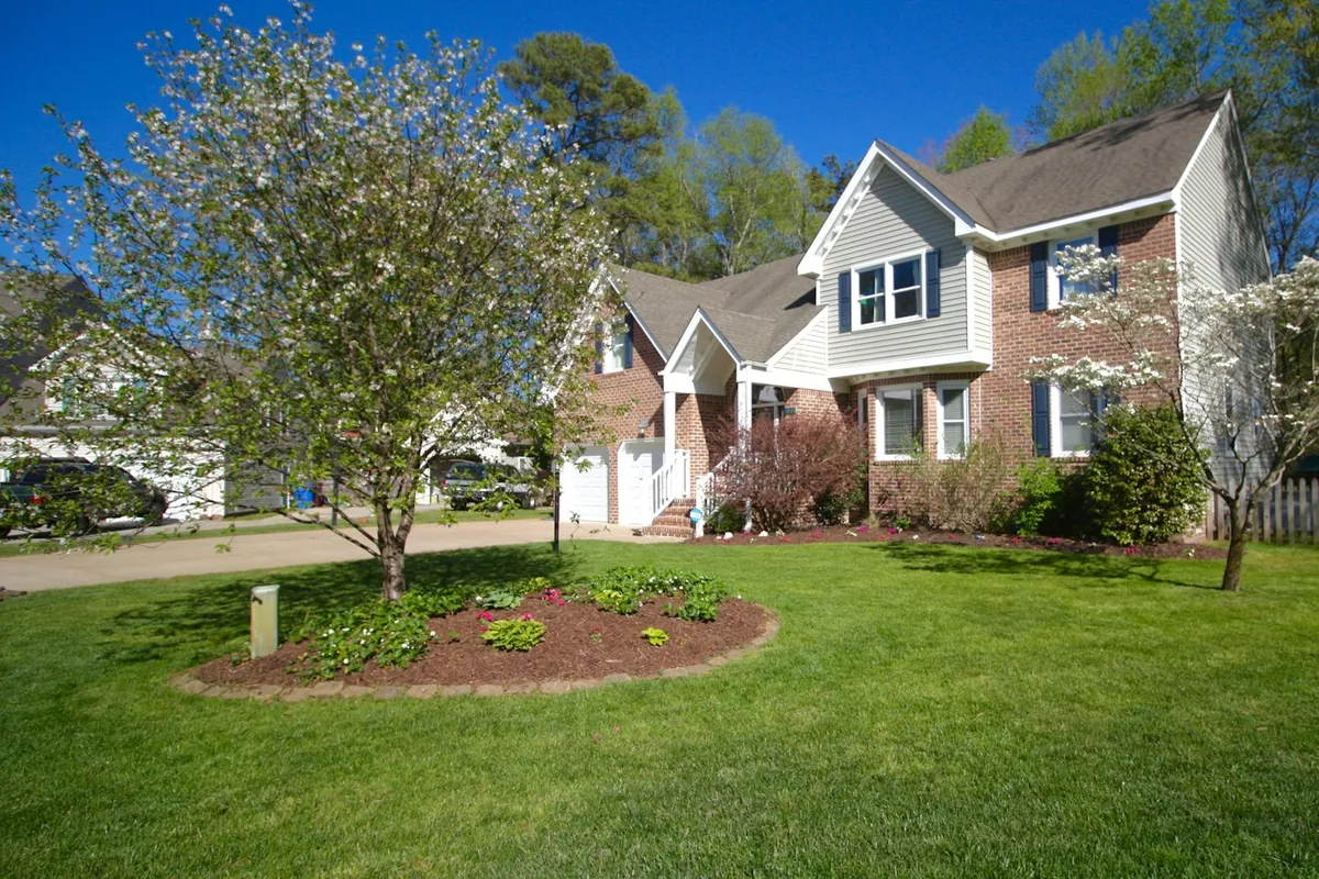 A young tree in a front lawn inside a round mulched bed, with a brick house behind