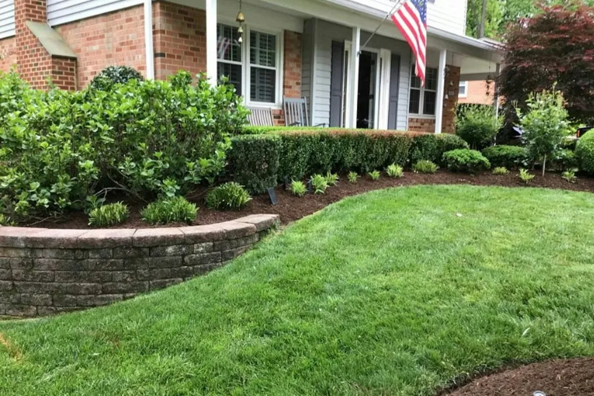 Mulched shrub beds along the front of a brick house, with a curved stone wall and a mown lawn