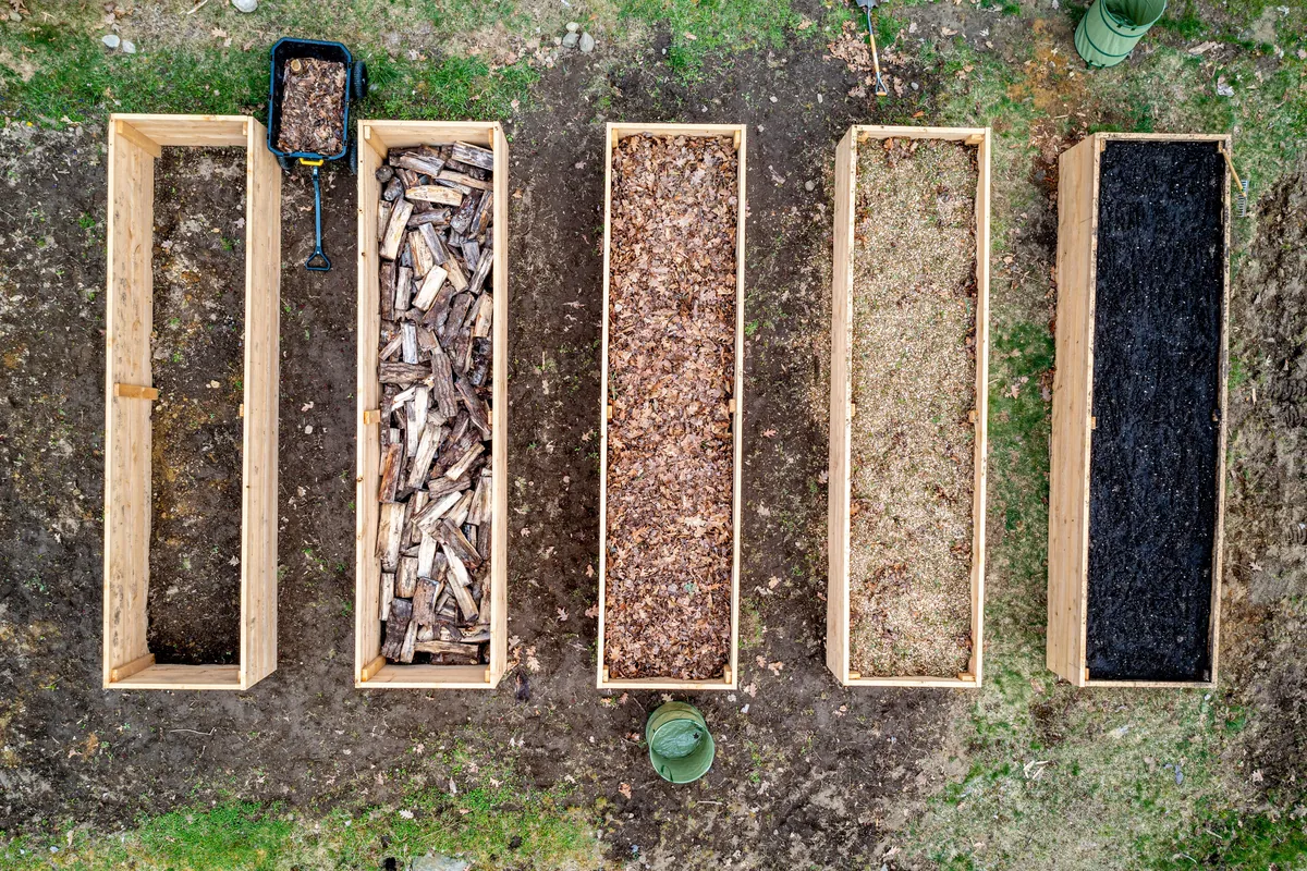Five new wooden bed frames seen from above, one empty and four holding logs, leaves, wood chips and a black fill