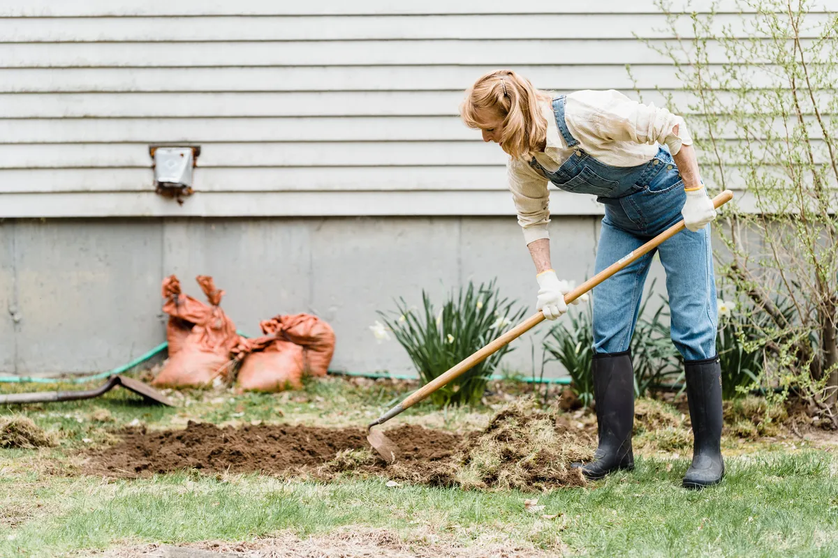 A woman in overalls hoeing up a strip of lawn beside a house, with sacks against the wall
