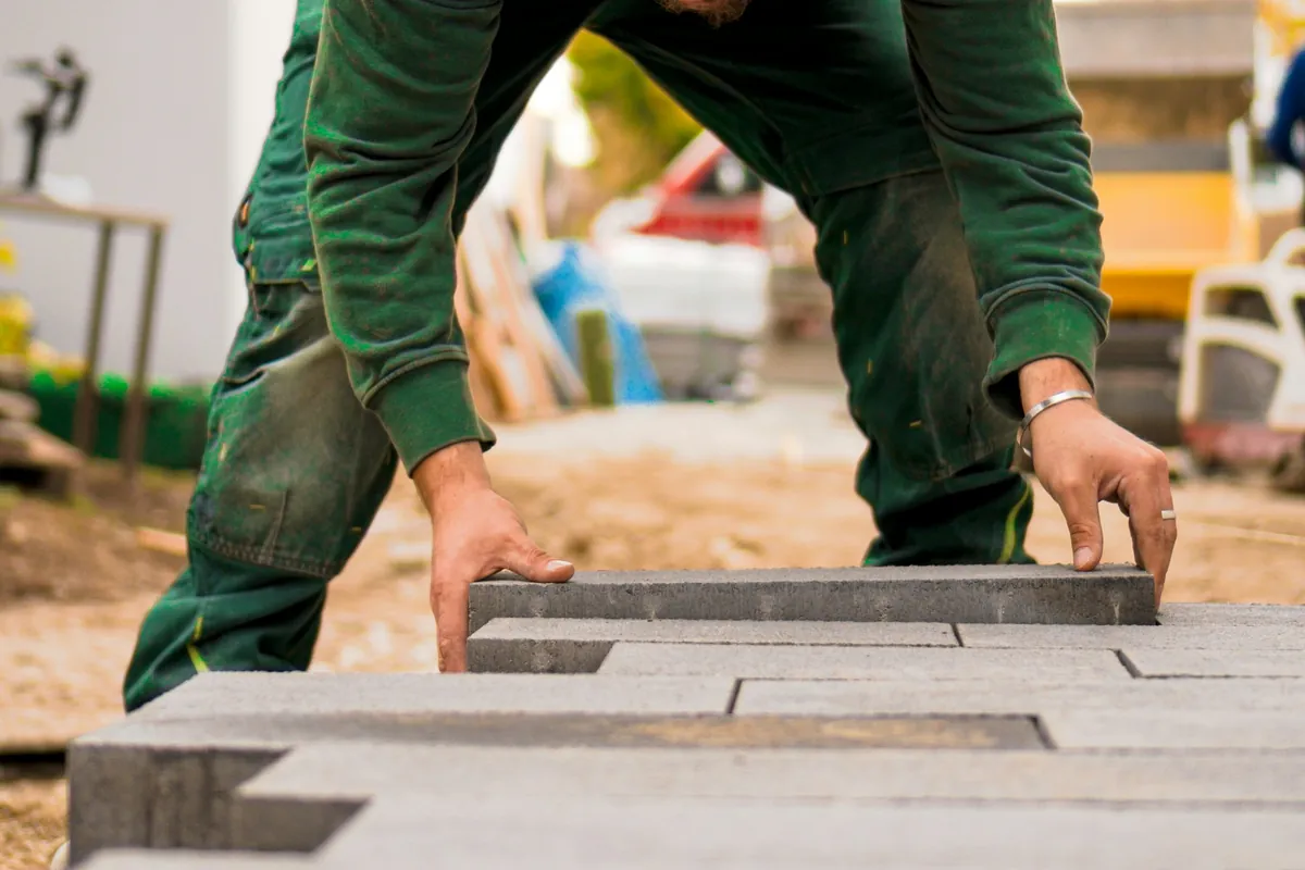 A worker in green work clothes setting concrete pavers on a sand bed